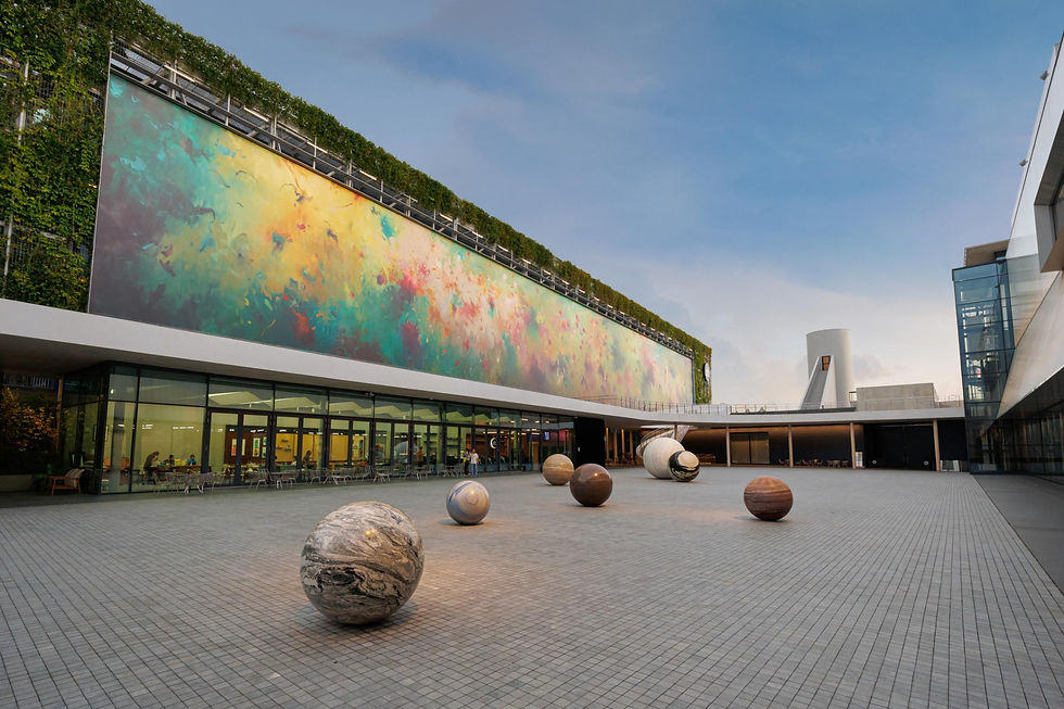 Modern courtyard with giant colorful mural, marble-like spheres on tiled plaza, and glass buildings under a clear sky
