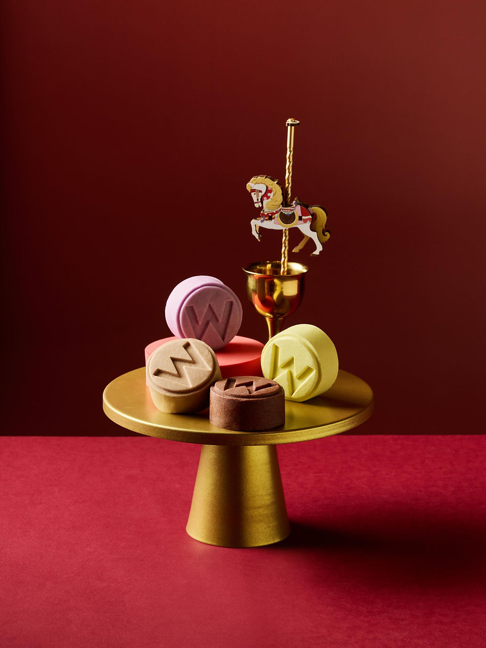 Colorful W-shaped pastries on a gold pedestal beside a tiny carousel horse, against a deep maroon backdrop.