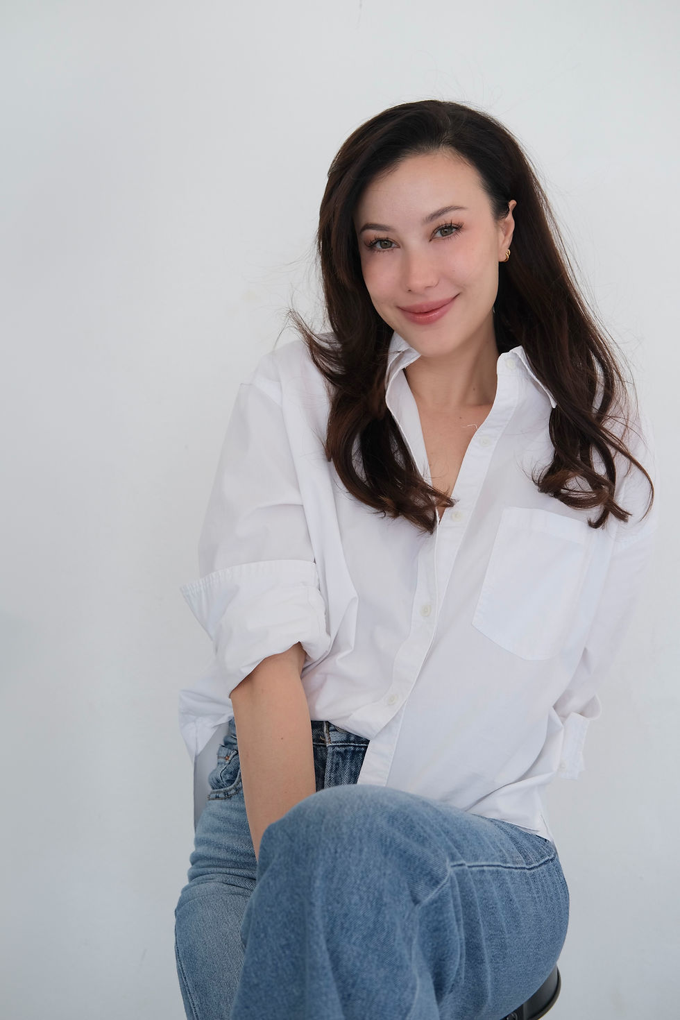 Woman smiling, seated against a plain white background. She wears a white shirt and blue jeans, exuding a relaxed, content mood.