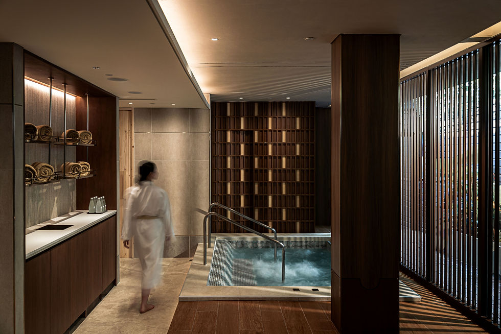 A person in a white robe walks towards an indoor hot tub. The spa setting features wooden panels, shelves with towels, and soft lighting.