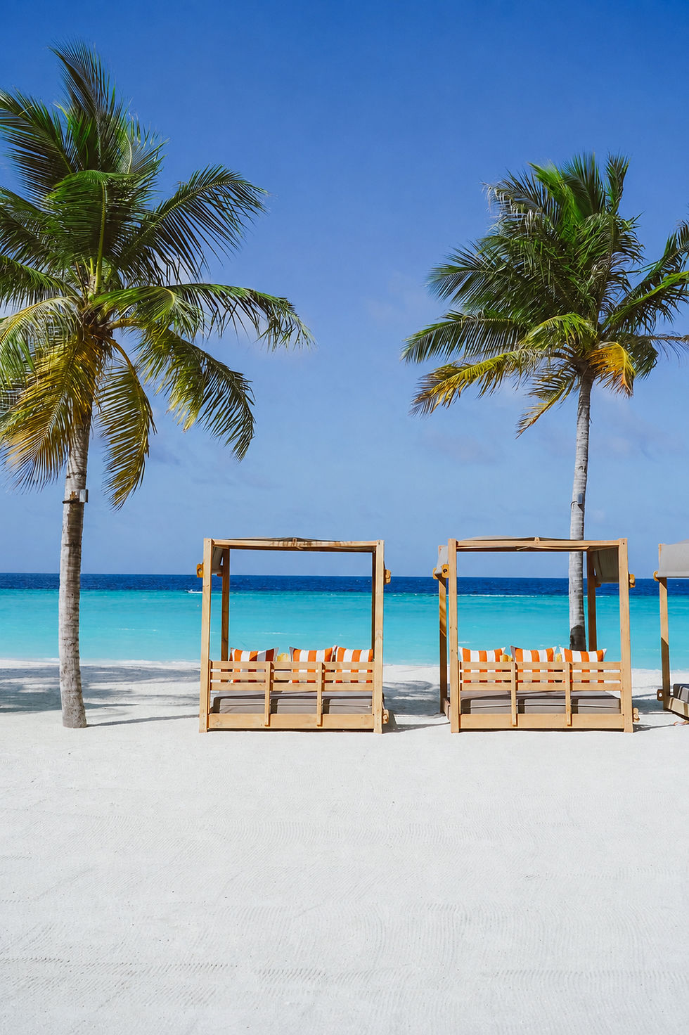 Palm trees frame empty beach cabanas on white sand beside turquoise water under a clear blue sky.