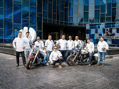 Chefs in white jackets pose confidently with motorcycles outside a modern blue building. Sign reads Renaissance. Bright, sunny day.