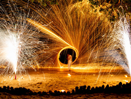 Fire performer in white amid orange and white spark trails at night, with audience silhouettes and trees overhead.
