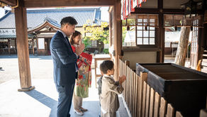 神社で礼拝する家族。父と子はスーツ、母は赤い着物。背景に伝統的な建物と青空が見える。穏やかな雰囲気。