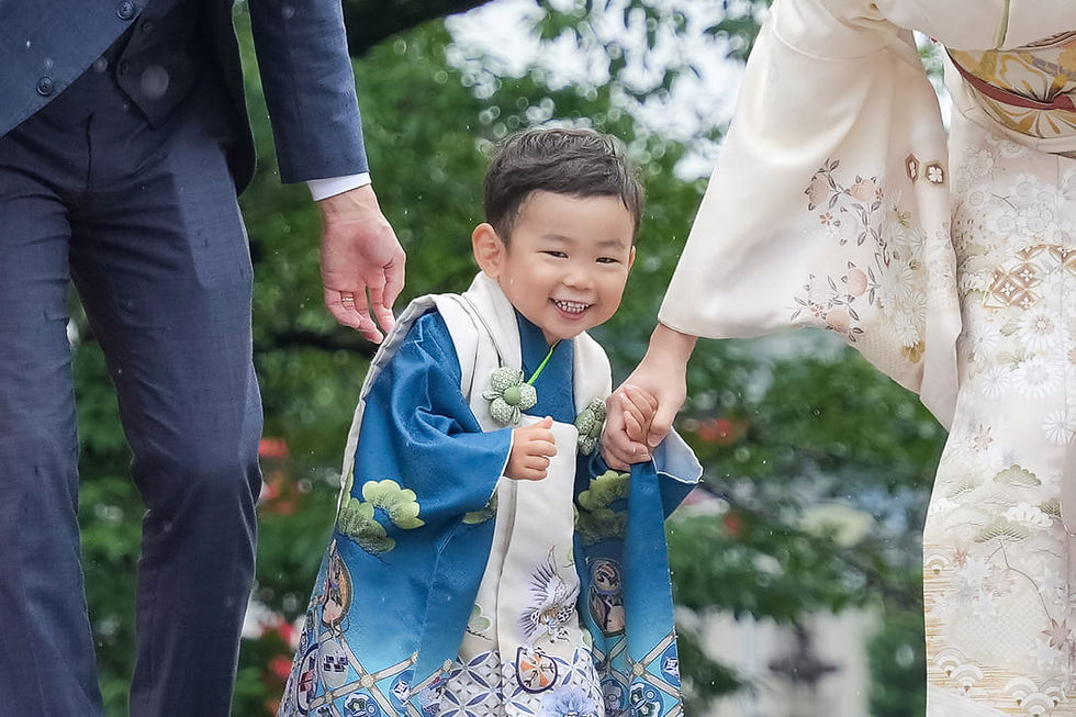 屋外で、青い着物姿の笑顔の幼い男の子が両親に手を引かれて歩いている。白い着物とスーツ姿、緑の木々が背景。