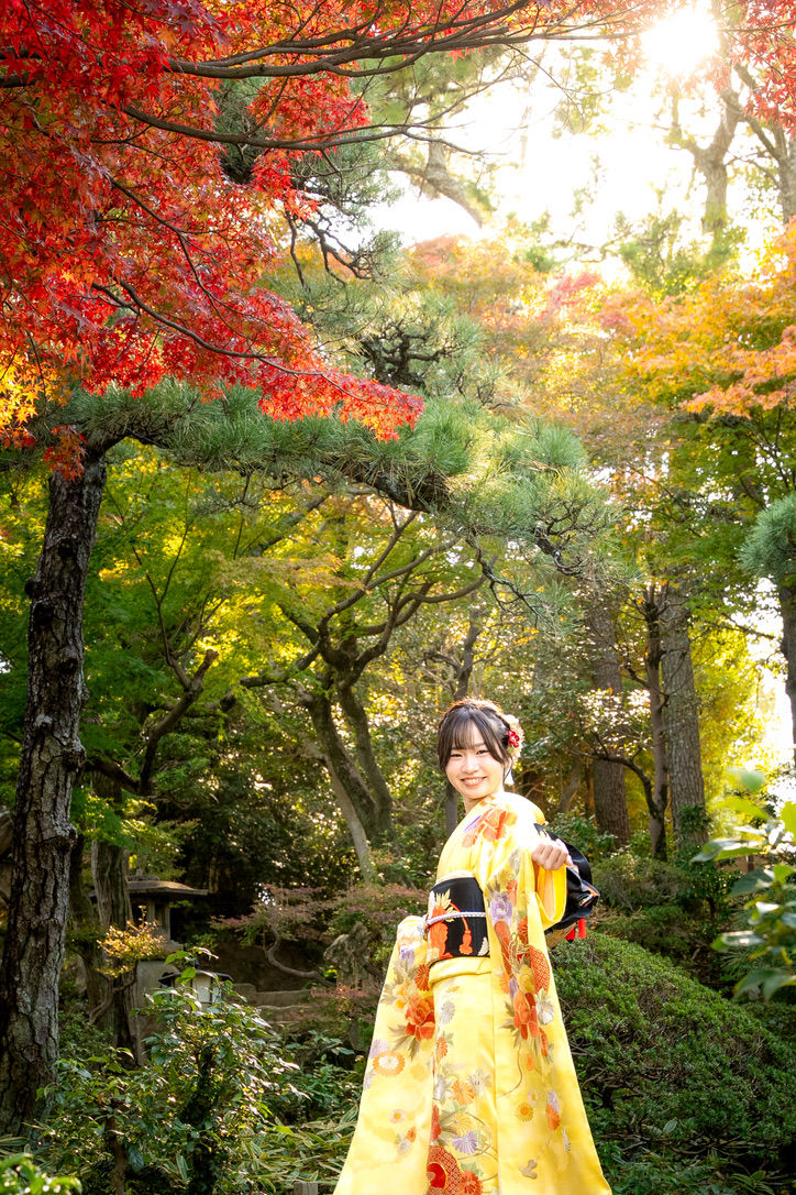 紅葉の庭園で、黄色の振袖を着た女性が笑顔で立つ。赤や緑の木々に陽光が差し込み、秋らしい明るい雰囲気。