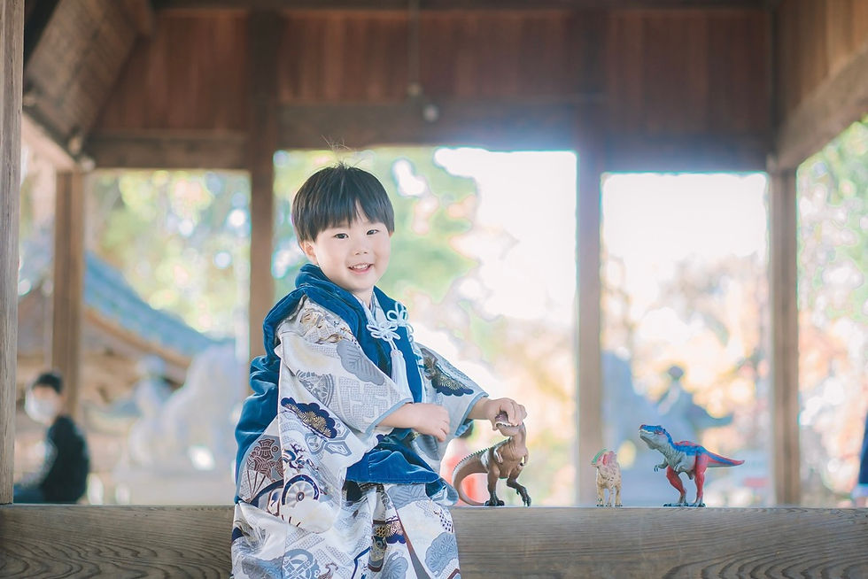 和服を着た男の子が寺院のベンチに座り、恐竜のおもちゃで遊んでいる。背景には木造建築とぼんやりとした秋の景色が広がる。