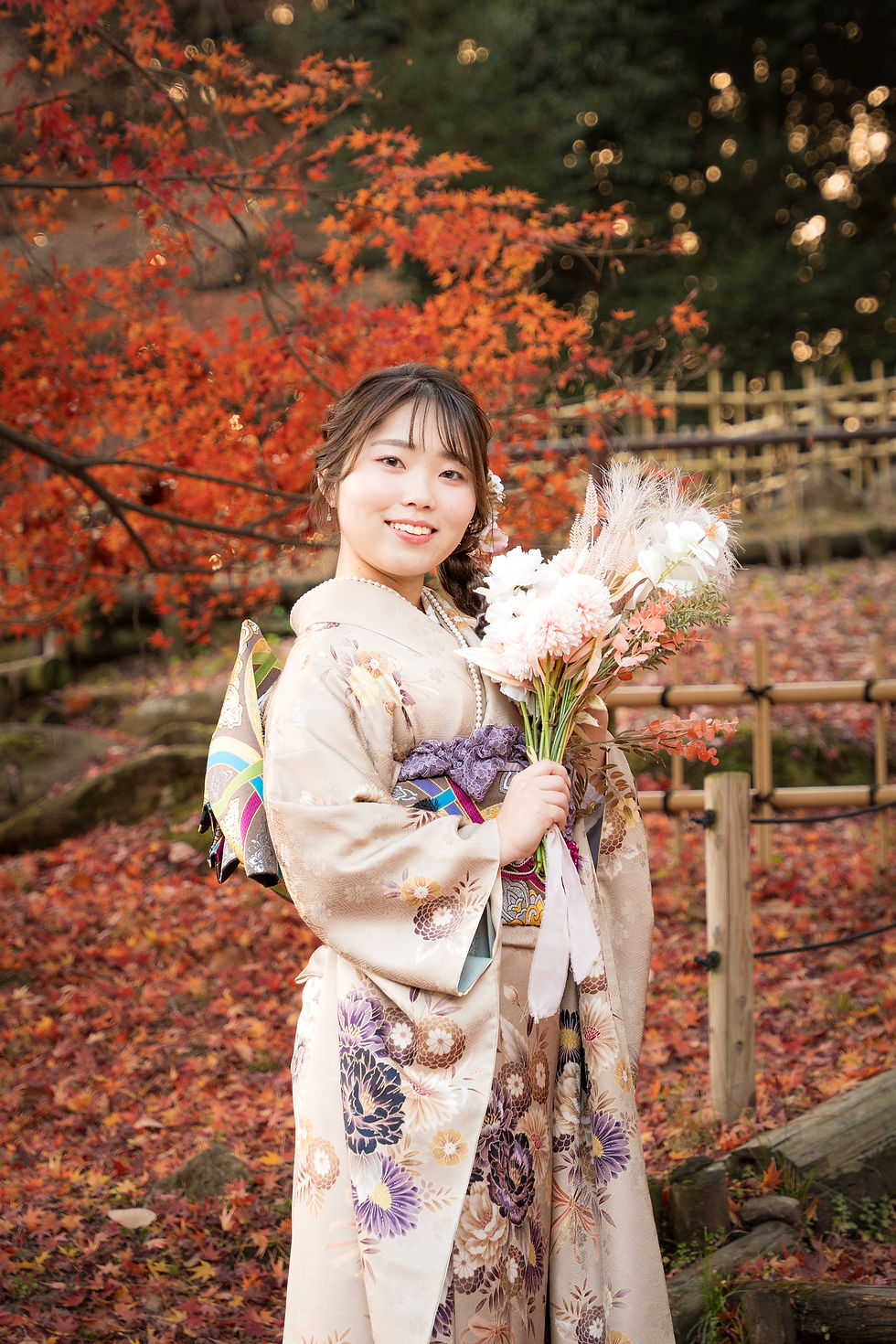 紅葉の庭で、淡い花柄の振袖を着た女性が花束を抱え、笑顔でこちらを見る秋の記念写真。