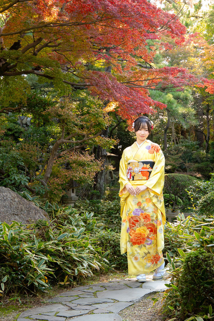 紅葉の庭園で、黄色い花柄の着物を着た女性が石畳の道に立ち、やわらかくほほ笑んでいる。