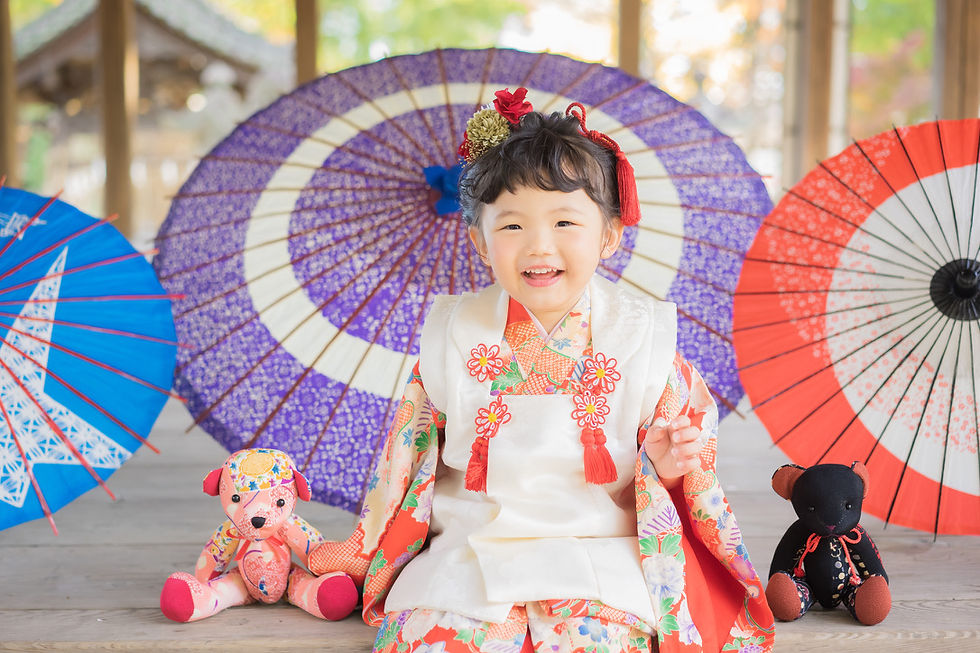 笑顔の幼い女の子が花柄の着物を着て座り、青・紫・赤の和傘とぬいぐるみに囲まれている。明るく華やかな雰囲気。