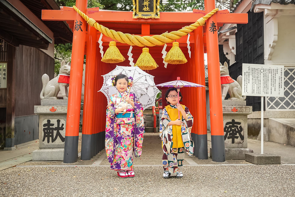 色鮮やかな着物を着た2人の子供が傘を持ち、神社の赤い鳥居の前に立っている。背景には狐の石像が見える。