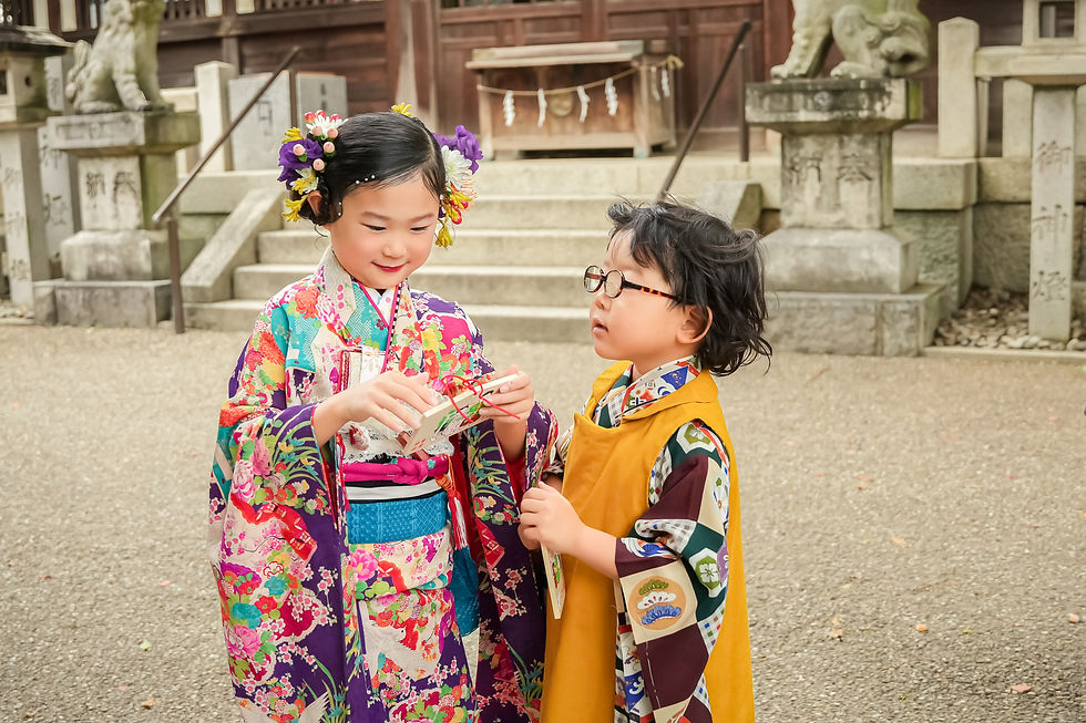 着物を着た二人の子供が神社の前で箱を持つ。女の子は華やかな花の飾りが特徴的で、男の子は眼鏡をかけている。穏やかな雰囲気。