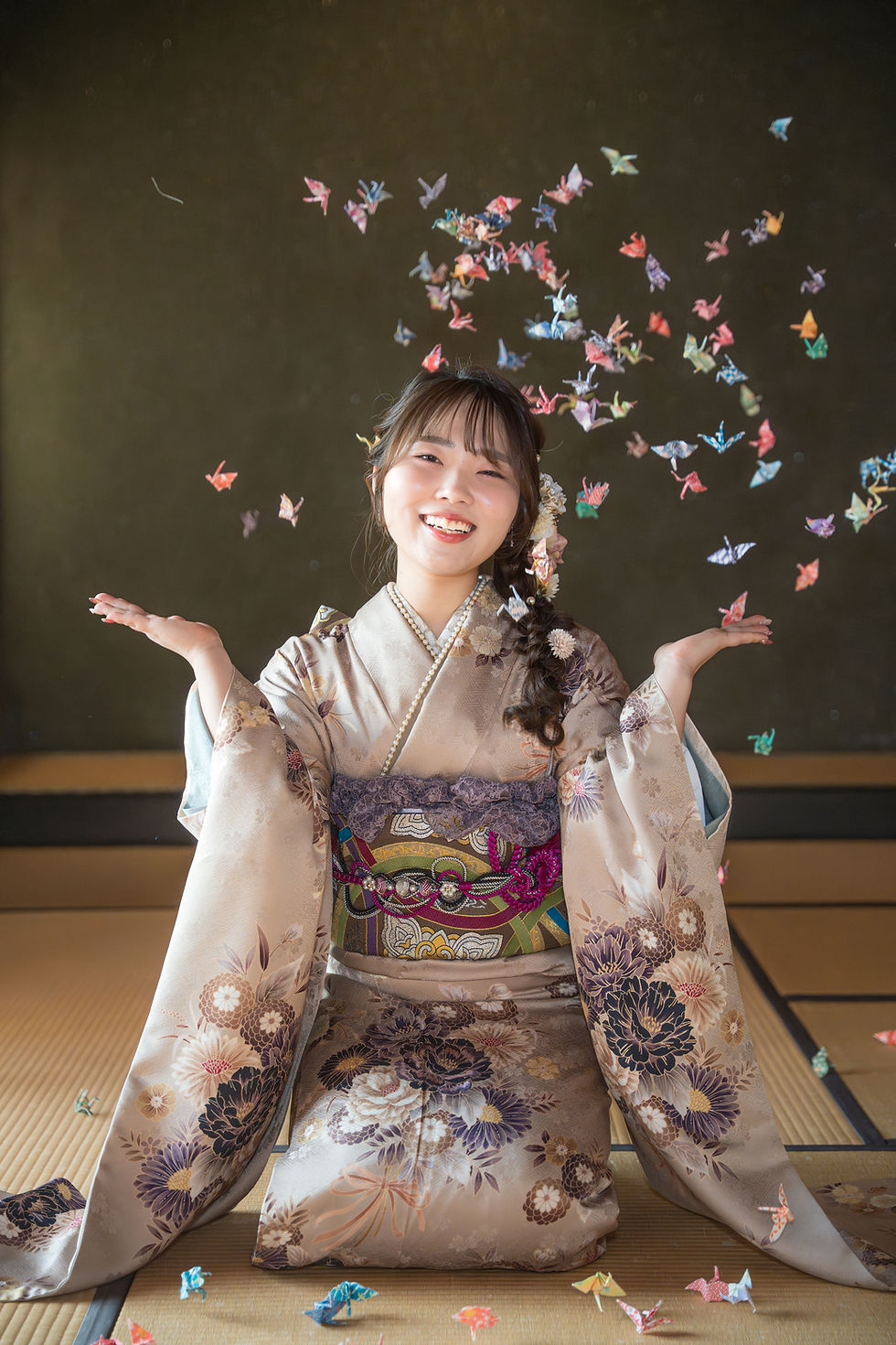 畳の和室で、花柄の着物を着た女性が笑顔で両手を広げ、周囲に色とりどりの折り鶴が舞っている。