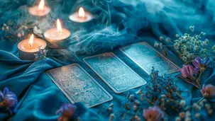 Três cartas de tarô dispostas sobre tecido azul, com velas acesas e flores ao redor, em cenário místico e espiritual.