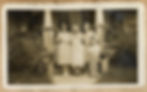 Three women stand side by side in front of a house's stone craftsman-style front porch. There is a young boy in striped overalls winking at the camera and standing in front of one of the women. 
