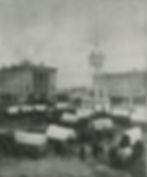 A black and white photo of a dozen covered wagons gathered around a monument in the center of the square. People and horses stand about. A stone building with large columns stands in the background to the left.