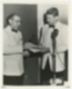 Lester E. Cox wearing glasses and a white tuxedo hands an award to Ronald Regan, in a matching outfit, as they stand in front of a microphone.