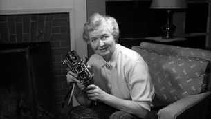 A woman sitting in an armchair in front of a fireplace holds a vintage camera