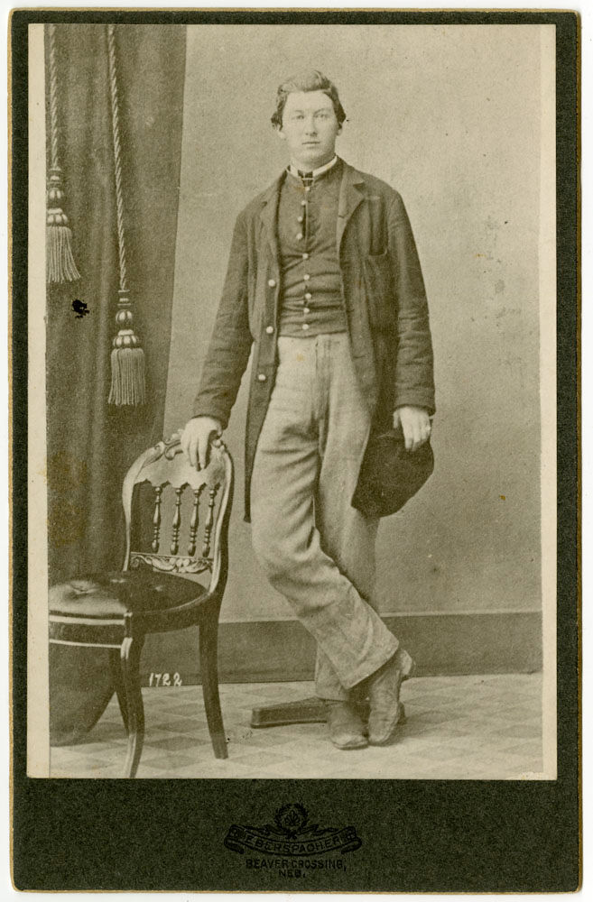 Black and white image of a man in turn of the century dress posing with his hand on the back of a chair.