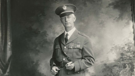 A man in a military style uniform stands in front of a painted backdrop