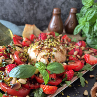 Burrata mit Erdbeeren, Tomaten, Pistazien und Basilikum auf Teller; im Hintergrund zwei dunkle Flaschen.