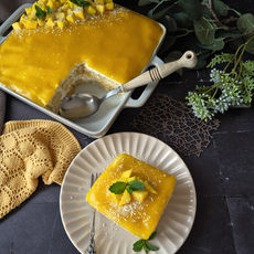 Mango-Dessert mit glänzend gelber Glasur auf Teller und Auflaufform, garniert mit Minze und Kokosraspeln, daneben Pflanzen und ein gelbes Tuch.