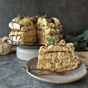 Ein Stück Schichttorte mit Schokostückchen liegt auf einem Teller. Die Torte mit Schokolade, Sahne und Minze dekoriert, im Hintergrund.