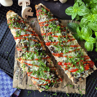 Zwei überbackene, mit Pilzen und Kräutern belegte Baguettes mit Ketchup auf Backpapier, daneben frischer Basilikum.
