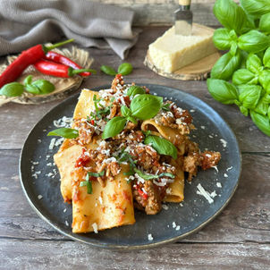 Pasta mit Fleischsauce, Basilikum und Parmesan auf schwarzem Teller, daneben rote Chilis und Basilikum auf rustikalem Tisch.