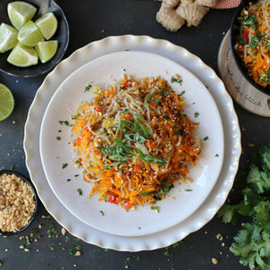 Bunter Glasnudel-Salat mit Karotten, Kräutern und Sesam auf weißem Teller, daneben Limetten, Nüsse und Ingwer auf dunklem Tisch.