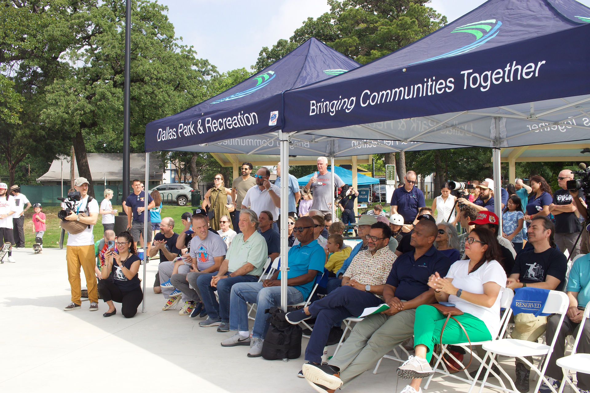 A view of the gathering crowd of guests, speakers and dignitaries under portable tents bearing the Dallas Park and Recreation logo and the words "Bringing Communities Together"
