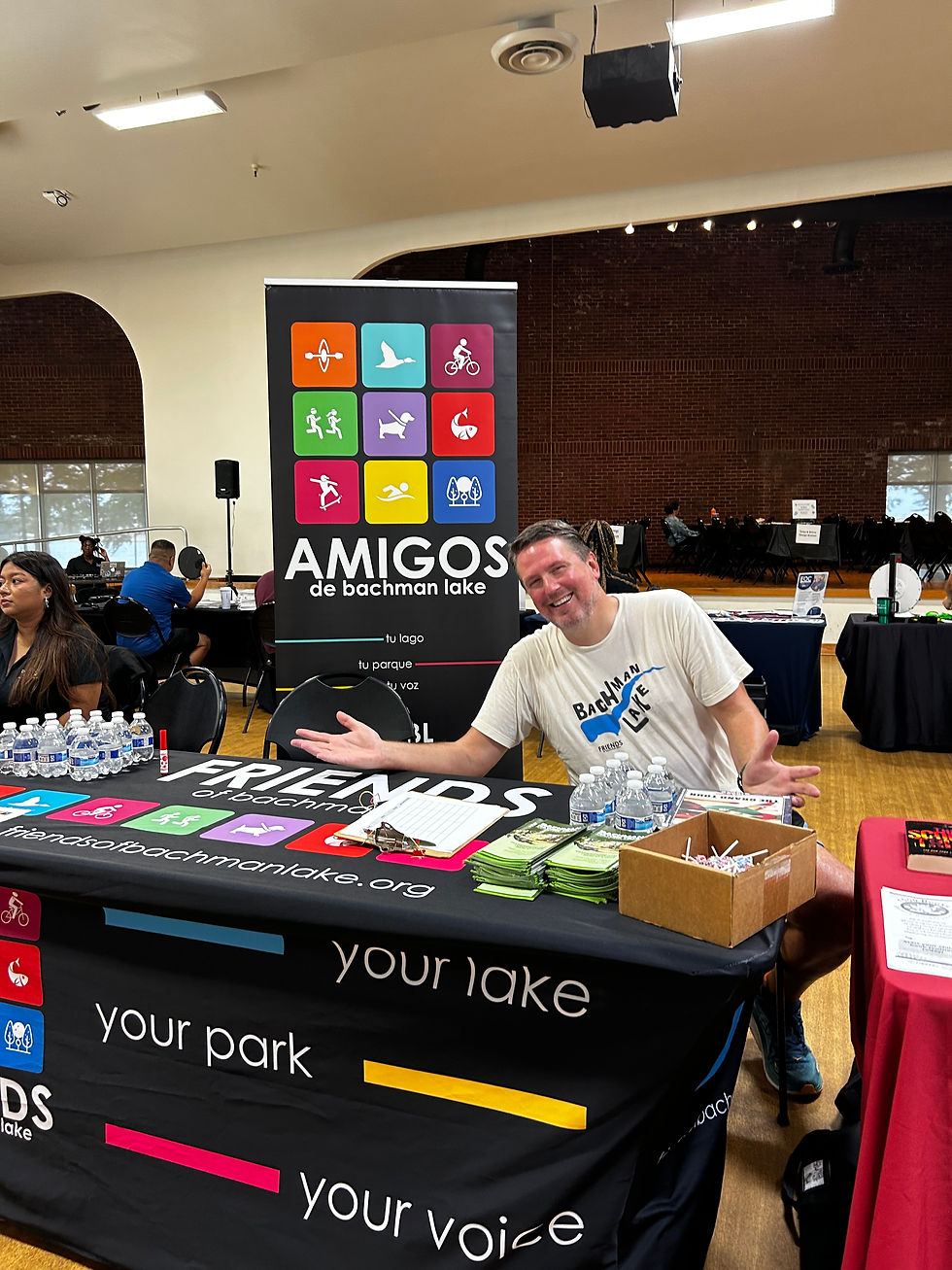 FoBL Board Member Jeff Brown welcomes attendees with Bachman flair at a table with a Friends of Bachman Lake themed table wrap, and a Amigos de Bachman Lake banner in the background.