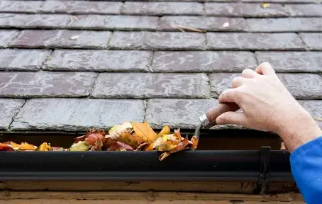 Hand cleaning leaves and debris from a roof gutter using a small tool.