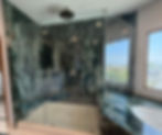Luxurious walk-in shower featuring glass doors, dark green marbled stone tile walls, and a overhead rainfall showerhead.
