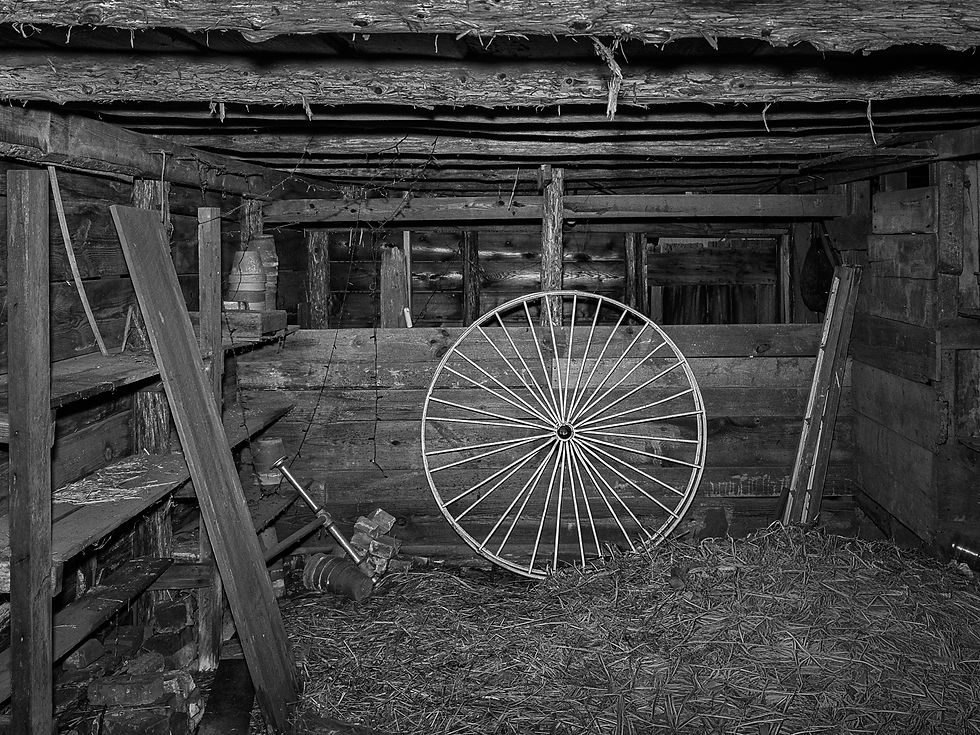 Black-and-white view of large spoked wheel and other objects inside an old barn.