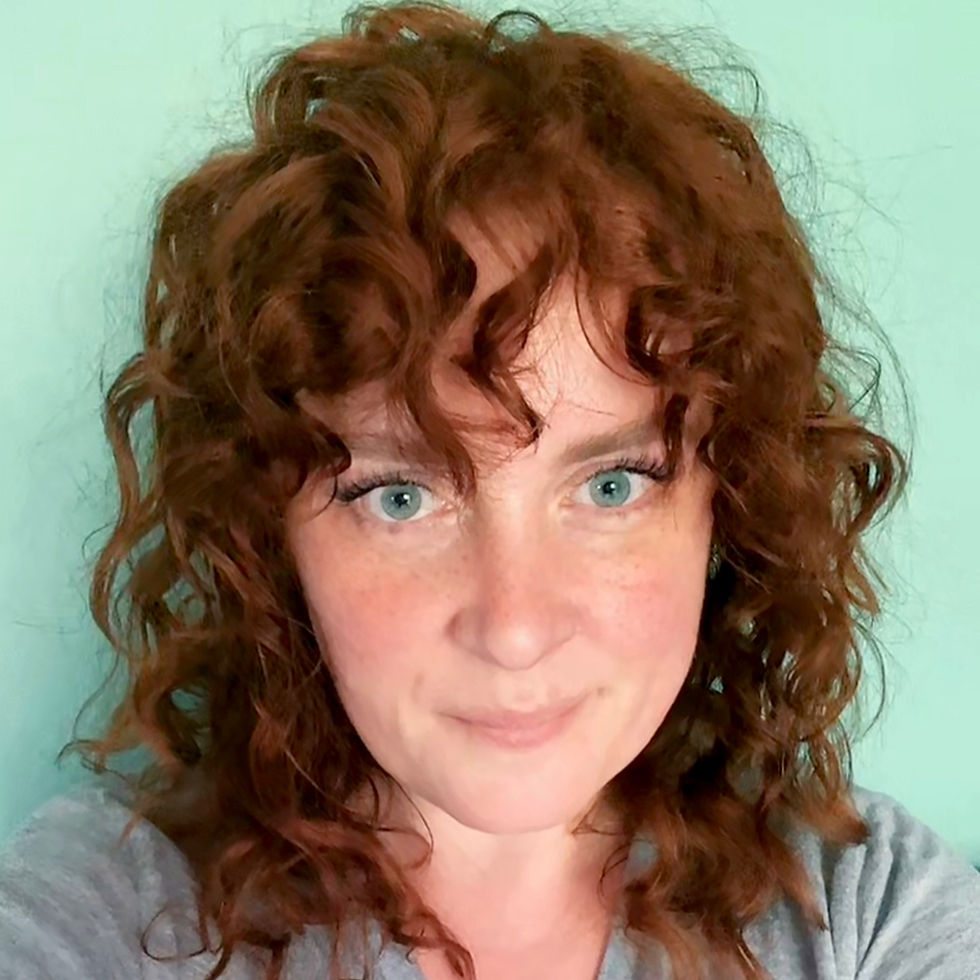 Woman with curly red hair and blue eyes smiles slightly against a light teal background, wearing a gray top. Mood is calm and relaxed.
