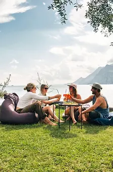 Vier Personen am Seeufer un Sitzsäcken stoßen mit Getränken an