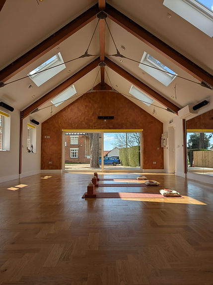 Interior view of Yoga at The Grange studio facing towards the patio windows. Three yoga mats are laid out on the wooden floor with bricks, blocks, blankets and eye pillows.
