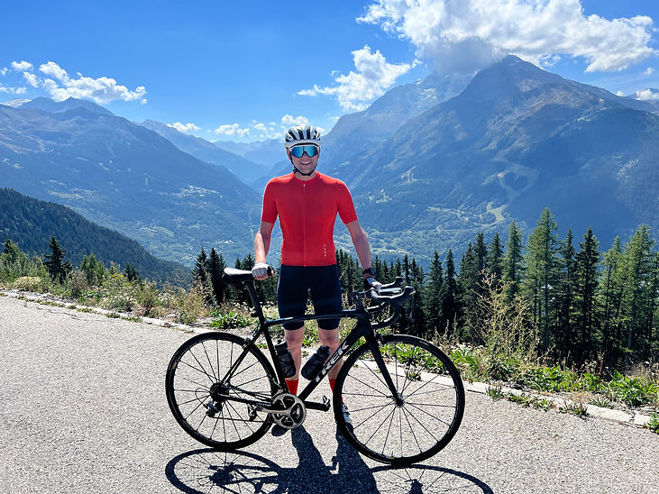 Adam holding his road bike infront of some pine trees and a beautiful mountain valley