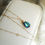 Thumbnail: Blue-green facted swarovski crystal gemstone on gold beaded chain, displayed on white leather jewellery box