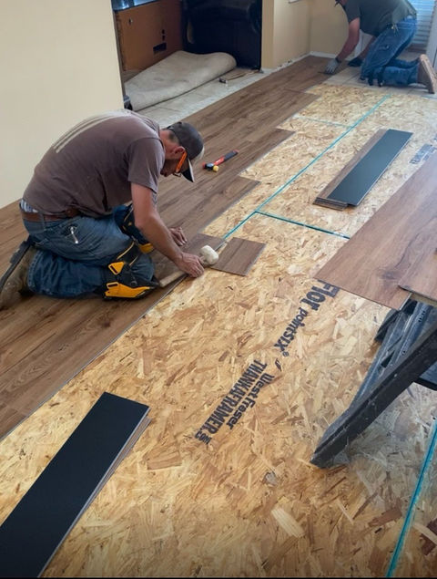 Man installing laminate flooring, using tools, on plywood subfloor in a room.