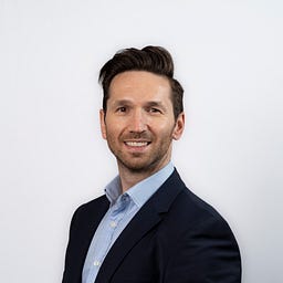 Smiling man with dark hair, wearing a blue shirt and suit jacket.