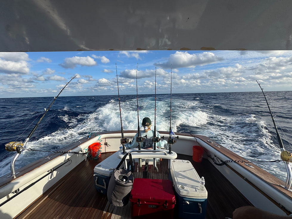 Fishing boat on open ocean with blue sky, white clouds. Five rods lined up, coolers and gear on deck. Wake visible behind boat.
