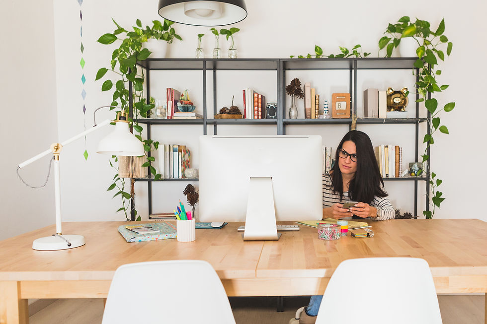 Woman in glasses using a smartphone at a desk. Shelves with books and plants in the background. Bright, organized workspace.