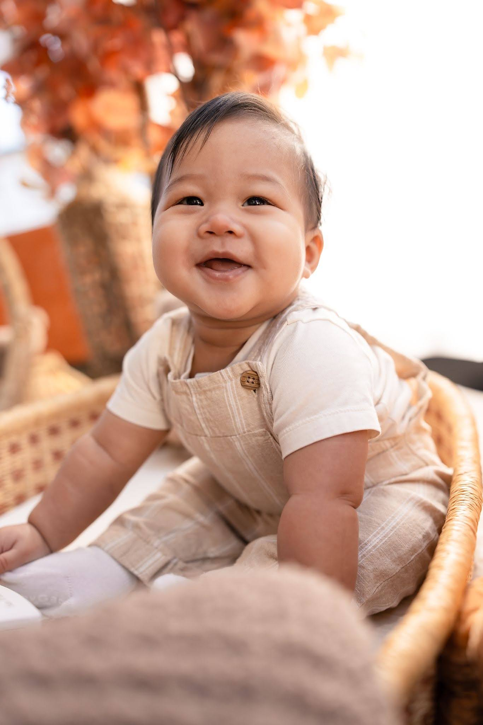 Smiling baby in beige overalls sitting in a wicker basket outdoors, with warm orange autumn leaves blurred behind.