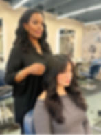 Stylist in black blouse adjusts woman's hair in salon. Woman wears gray top, looks serene. Modern decor in background.