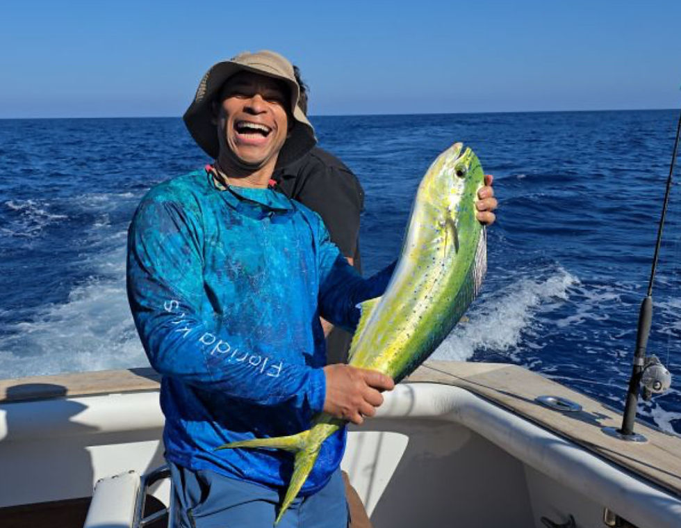 Man in a blue shirt and hat joyfully holds a large green fish on a boat with the ocean in the background. Bright, sunny day.