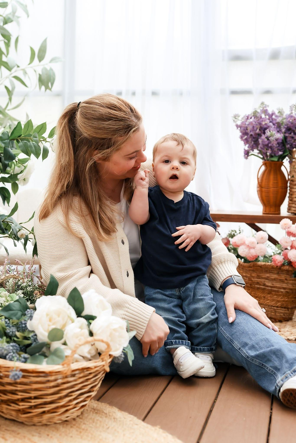 Woman holding baby in a bright flower-filled room, gazing at him while he looks at the camera.