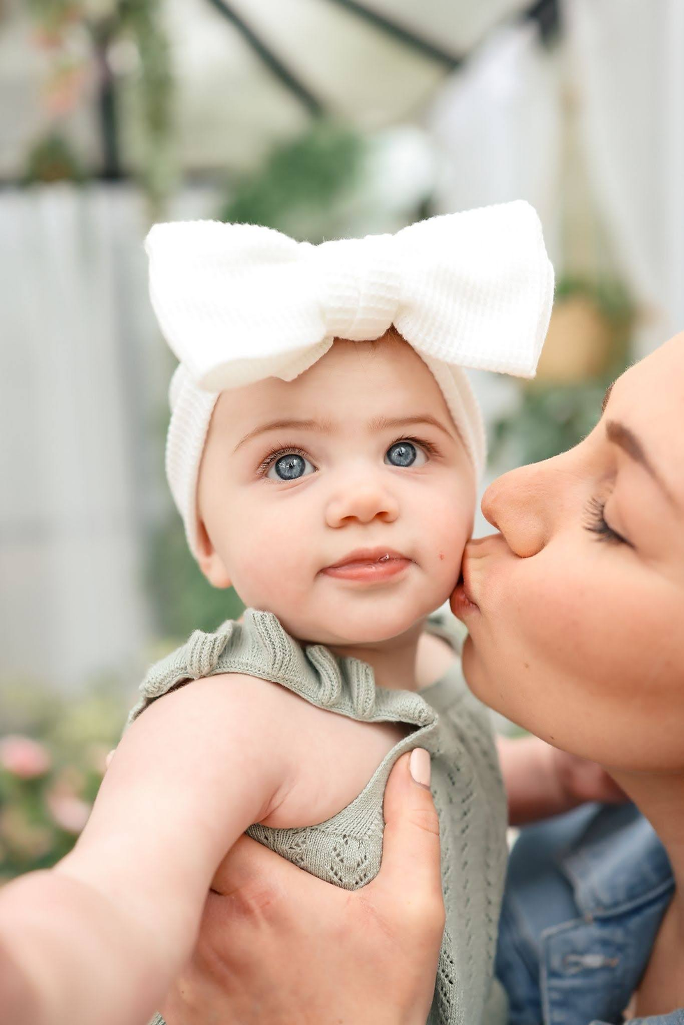 Woman kissing a baby’s cheek; baby in a white bow headband and green outfit looks at the camera, with a soft blurred background.