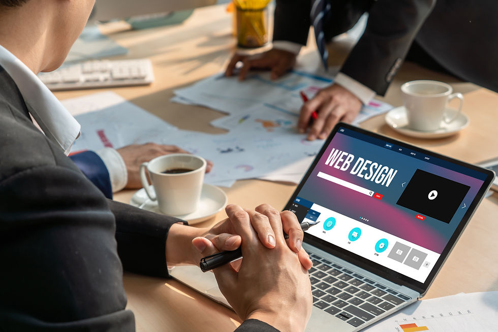 Person in suit views a "Web Design" screen on laptop. Papers, coffee, and colleagues in the background suggest an office meeting.